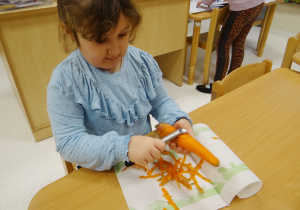Zuzia siedzi przy stoliku i obiera marchewkę.